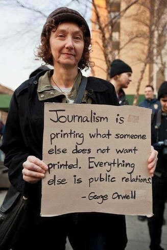 Woman holding up a sign on cardboard with a George Orwell quote about the nature of journalism vs public relations.