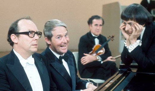 Morecambe & Wise at the piano whilst an exasperated Andre Preview looks on, his head in his hands.