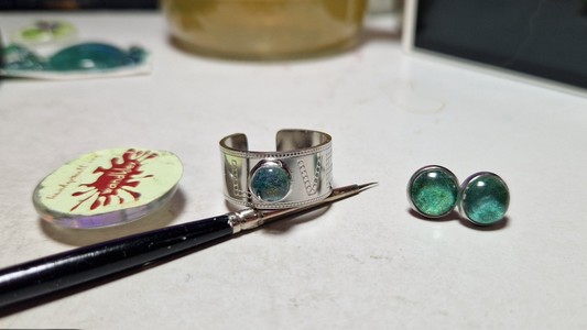 Silberfarbener Edelstahlring mit Gravur love, das O ist eine Fassung für ein rundes handgemaltes galaxienmotiv in grün,daneben liegen Ohrstecker in derselben Farbe und ein Pinsel zur Verdeutlichung dass es sich um handgemalte Stücke handelt auf den nächsten Fotos ist das gleiche nur aus einer anderen Perspektive bzw. an der Hand fotografiert zu sehen