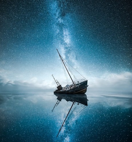A shipwrecked boat leans on its right side in calm water. Both the boat and the incredibly starry sky above reflect perfectly in the water's mirror like surface.