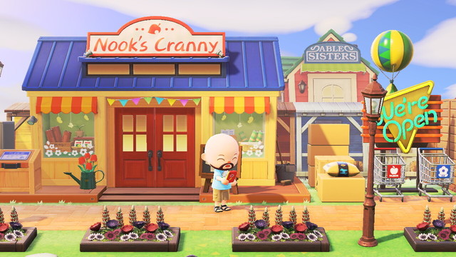 Animal Crossing: New Horizons scree shot. A bald, bearded, light skinned villager is looking at a red smartphone right outside of Nook's Cranny. On the right of the building are various piles of boxes, a "We're Open" sign, and two shopping carts. There are flower beds in front of a brick walkway that goes in front of the shop. The Able Sisters shop can be seen in the background.
