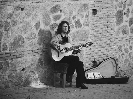 Flamenco busker sitting on a stool next to an old stone wall playing his guitar. His case is nearby with CDs (?) and to receive tips. He is dressed the part with longish hair. He wears a serious expression.