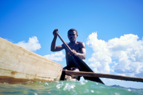African man paddling a wooden canoe. Photo is taken from water level on the side of the canoe. Blue sky with white clouds in the background.