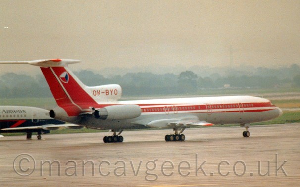 Side view of 3 engined jet airliner with the engines mounted on the rear fuselage, taxiing from left to right and slightly away from the camera.The plane is white, with a grey belly, and a pair of red stripes running along the body, getting slightly wider as they go, until they sweep upo in to the tail.
The registration "OK-BYO" is on the centre engine intake in red.
There is a logo on the tail that looks a little bit like a squashed cube, with red, white, and blue faces.
In the background, there is a grey and blue jet airliner just behind this planes tail, while grass runs off into the distance, trees and hills beyond the airfield perimeter getting lost in haze.
