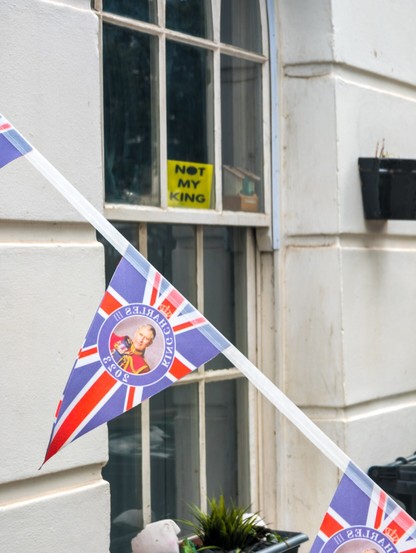 Cropped view of a house window in a white wall, with a yellow 'NOT MY KING' sign prominently visible. King Charles III 2023 Union Jack bunting crosses in front of the shot