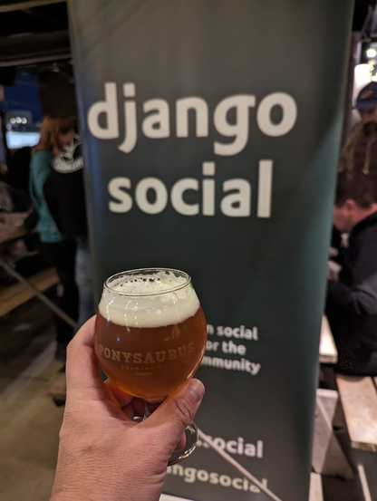 A snifter glass full of a fresh hop IPA held up in front of the Django Social banner