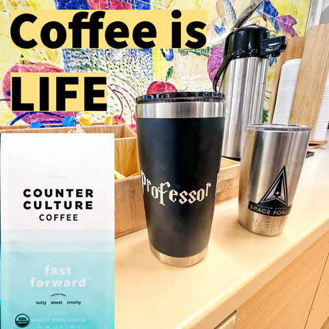 A coffee-themed setup featuring a bag of Counter Culture Coffee labeled "fast forward," a black travel mug with "Professor" written on it, a stainless steel tumbler with a Space Force logo, and a coffee carafe, all against a colorful mural