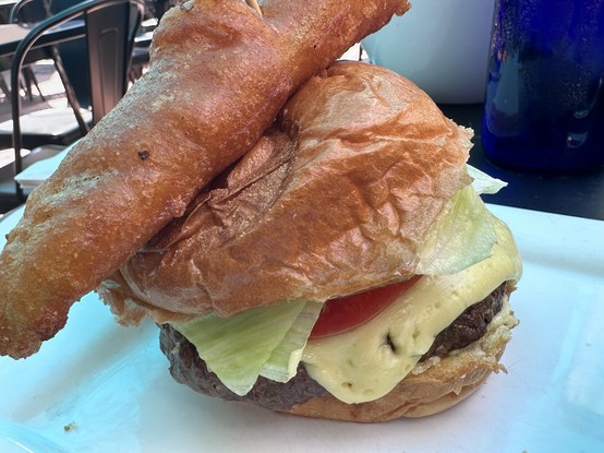 A close-up of a hamburger featuring a golden-brown bun, a beef patty, melted cheese, lettuce, and tomato. A crispy fried pickle is placed on top of the bun. The background consists of outdoor dining elements.