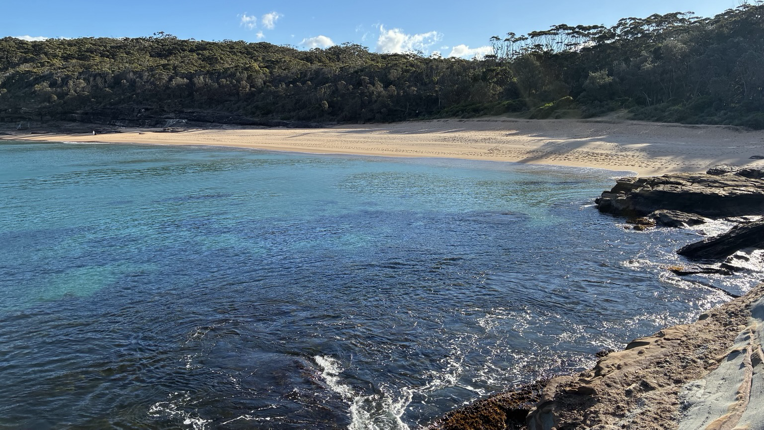 Emily Miller Beach from the headland an Arc of white sand and bush behind with blue green Stillwater and rocks in the front